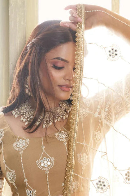 Woman in traditional beige embroidered outfit with jewelry, standing by a window.